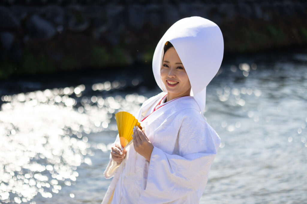 白無垢姿の新婦が屋外で扇子を手に立つ神前式の記念写真。背景には水面の輝きが広がる。