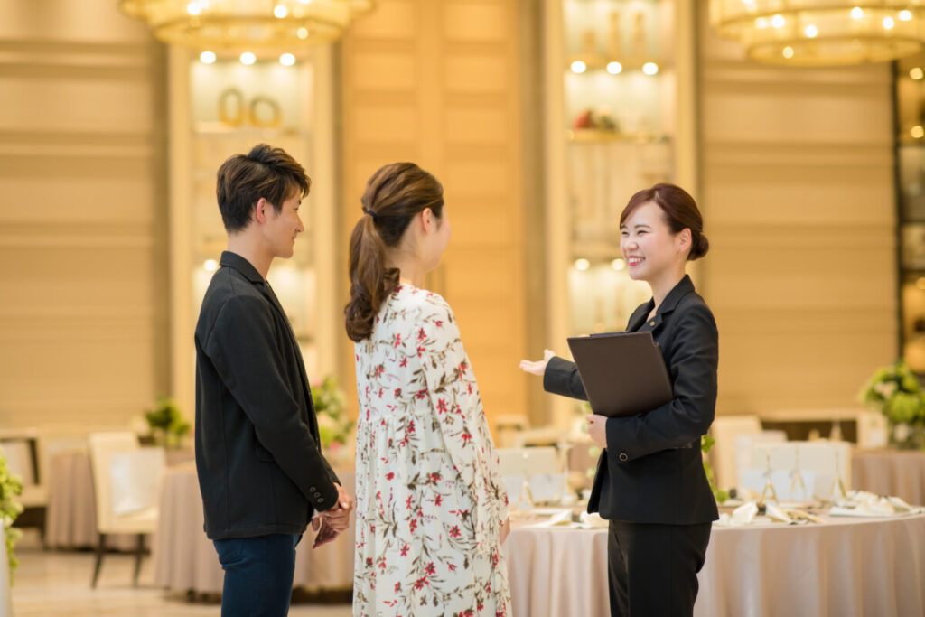 Luxscena（ラグシエナ／三島・沼津・富士エリア）の披露宴会場で、プランナーが新郎新婦へ結婚式の会場見学と相談案内を行っている様子。ブライダルフェア（BRIDALFair）で体験できる上質な空間が伝わるシーン。