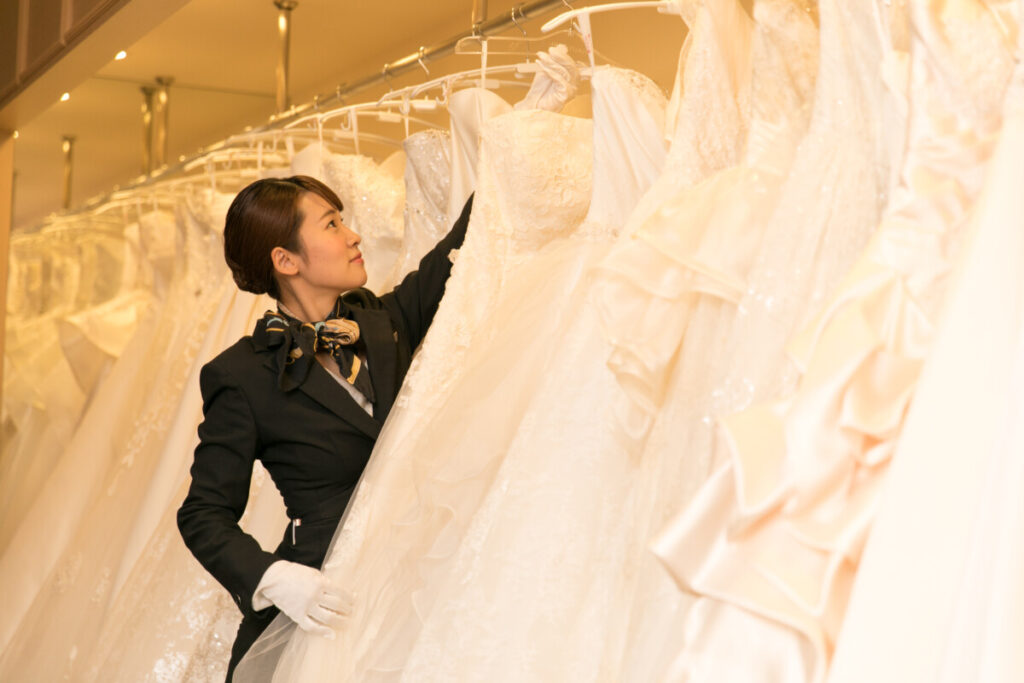 三島・沼津・富士エリアでフォトウェディングや前撮りを検討する新郎新婦の衣装合わせシーン。専属スタッフがウェディングドレスを丁寧に案内するロケーションフォトの準備風景。