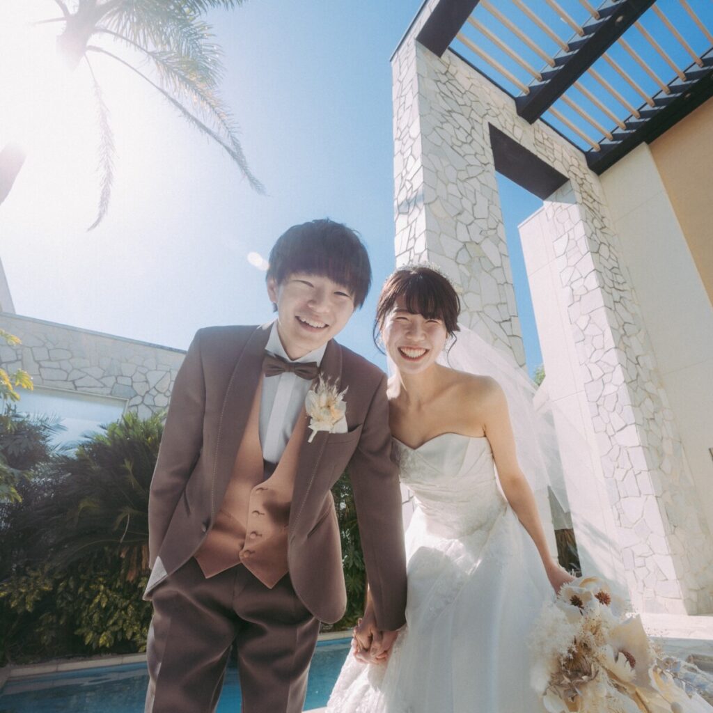 三島・沼津・富士エリアでフォトウェディングや前撮りを検討する新郎新婦のロケーションフォト。青空と水辺、緑に囲まれたリゾート空間で手を取り合う洋装の撮影シーン。
