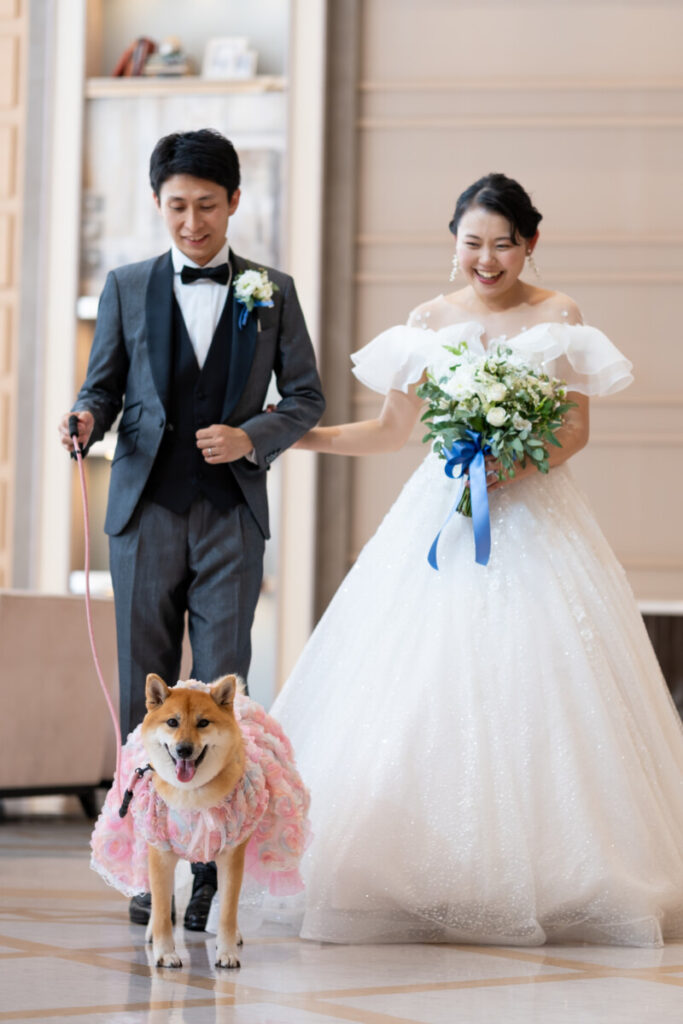 ラグシエナのペットウェディングで、新郎新婦と愛犬（柴犬）が並ぶフォトシーン。ピンクのドレスを着た愛犬と、ブーケを持つ新婦、リードを持つ新郎が室内で撮影した温かな家族ショット。