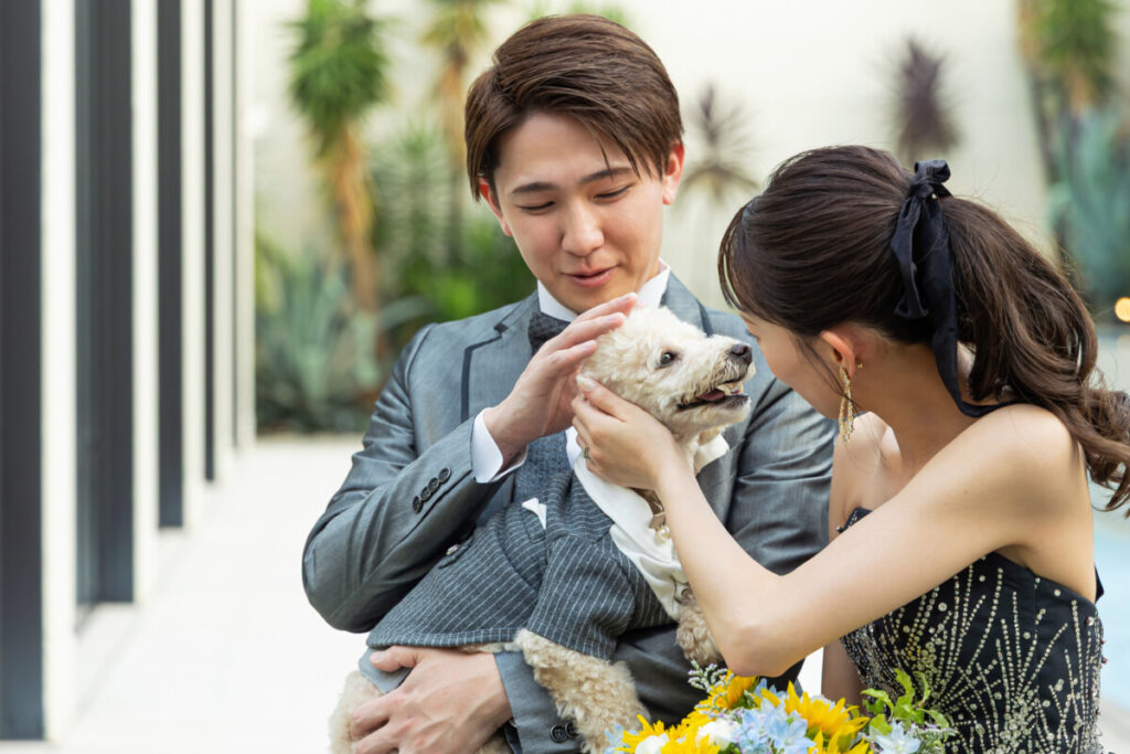 ラグシエナのペットウェディングで、新郎新婦がフォーマル衣装の小型犬と触れ合うガーデンフォト。愛犬が主役になる結婚式の温かなワンシーン。