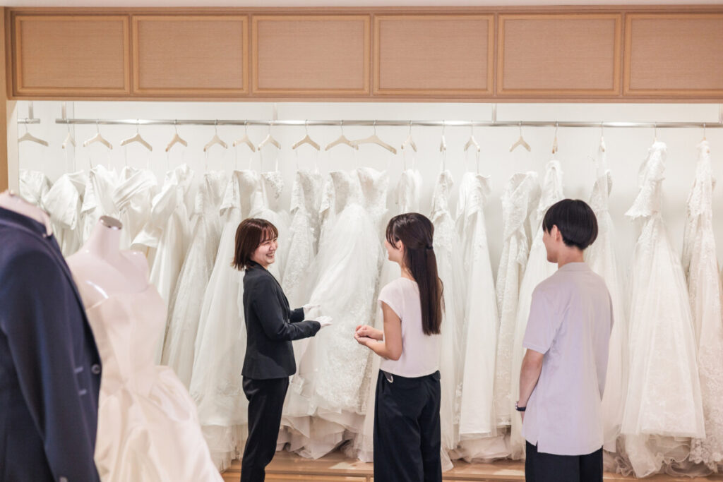 Luxscena（ラグシエナ／三島・沼津・富士エリア）のブライダルフェアで、プランナーが新郎新婦へ結婚式・披露宴の衣装見学を案内している様子。ウェディングドレスが並ぶ上質な空間での相談シーン。